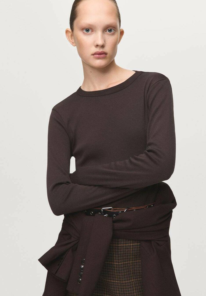Tmavohnedý top s dlhým rukávom a zaobleným výstrihom, vyrobený z mäkkej látky. Doplnkovaný uzlovým detailom v oblasti pásu, navrstvený cez kárované nohavice.
