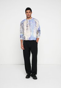 Miesten neulepaita, jossa on sinivalkoinen abstrakti kuvio, pyöreä pääntie ja pitkät hihat, yhdistetty mustiin laskostettuihin housuihin ja kenkiin.