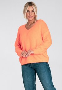 Kobieta z blond włosami, ubrana w jasnopomarańczowy sweter i niebieskie dżinsy, stojąca na tle jasnego, jednolitego tła.