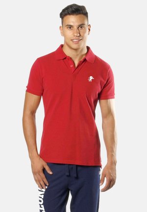 Homme portant un polo rouge avec emblème de lion et un pantalon de survêtement bleu marine avec texte blanc, debout devant un fond uni.