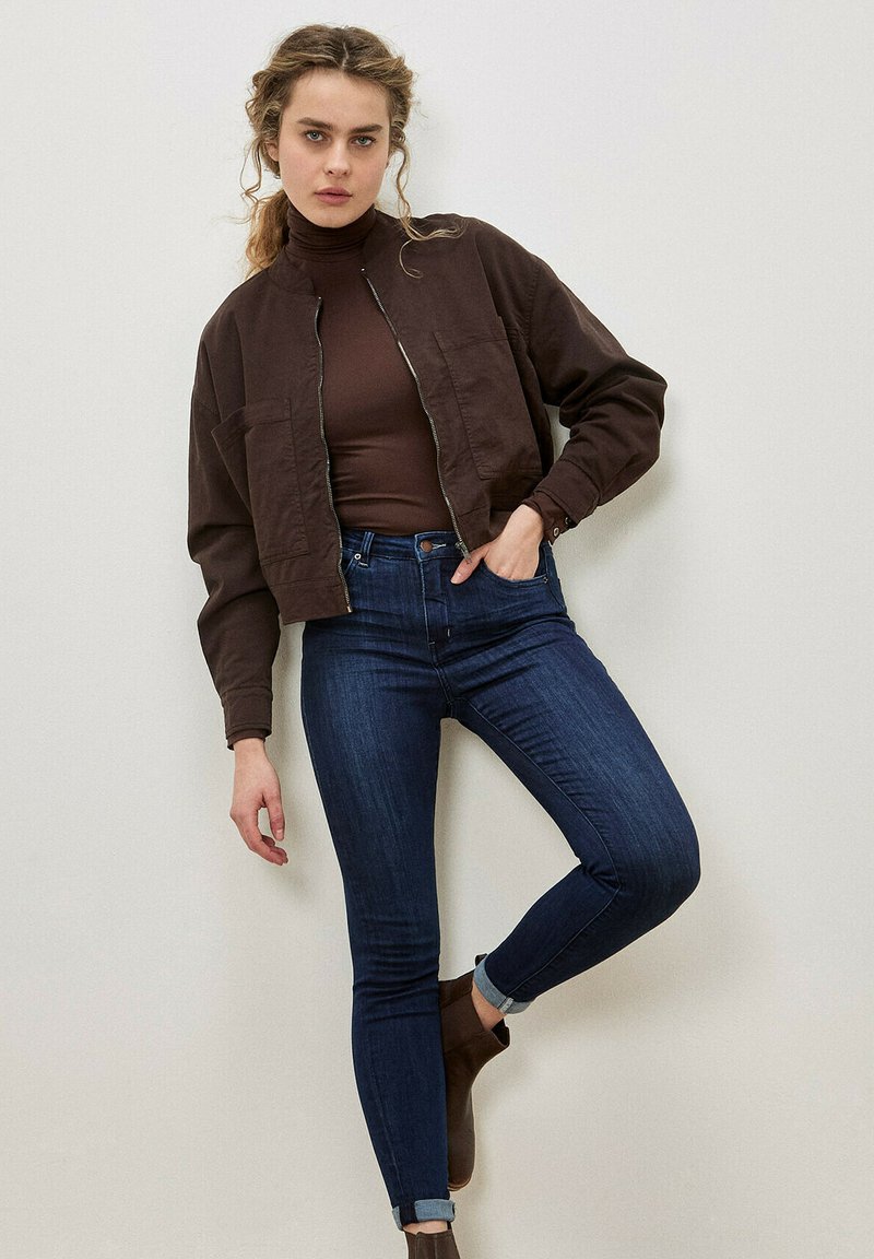 Chaqueta corta marrón con dos bolsillos frontales sobre un jersey de cuello alto marrón ajustado, combinada con vaqueros azul oscuro y botas de tobillo marrones.
