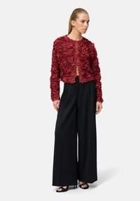 Veste texturée à motifs floraux rouges avec un col rond, devant ouvert, portée sur un pantalon noir à jambes larges et des sandales à talons. Fini en tissu lisse.