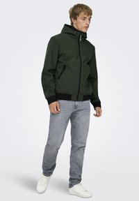 Giacca con cappuccio di colore verde scuro, realizzata in tessuto liscio, con zip frontale, due tasche laterali e polsini e orlo a coste. Indossata con jeans grigi.