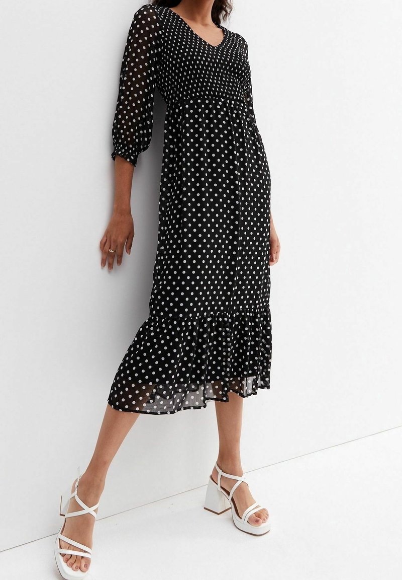 Schwarzes Kleid mit weißen Punkten, mit dreiviertellangen, transparenten Ärmeln, V-Ausschnitt und Rüschensaum. Kombiniert mit weißen Blockabsatzsandalen.