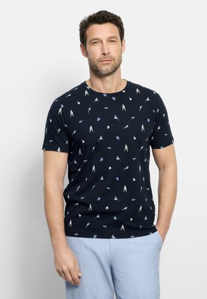 Uomo che indossa una maglietta blu navy con stampa a tema tennis e pantaloni azzurri, in piedi davanti a uno sfondo chiaro e uniforme.