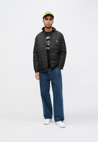 Svart quiltet jakke med trykknapper, brukt over en svart t-skjorte med grafisk trykk. Kombinert med løse blå jeans og hvite joggesko.