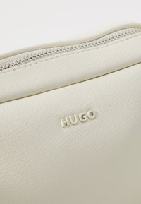 White textured leather bag with a silver zip. Features embossed "HUGO" logo on the front, highlighting minimalist design and craftsmanship.