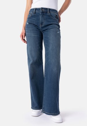 Donna che indossa jeans a vita alta blu scuro a gamba larga con lievi dettagli di usura e sneakers bianche, in piedi su uno sfondo neutro.