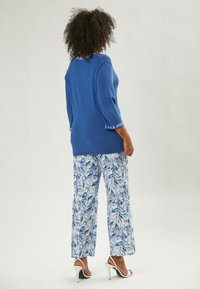 Top blu in maglia con maniche a tre quarti e orlo decorato, abbinato a pantaloni bianchi con stampe di foglie blu. Sandali bianchi con tacco alto.