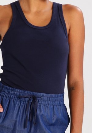 Femme portant un débardeur sans manches bleu marine et un pantalon à taille élastique avec un cordon de serrage bleu, avec un tatouage partiel visible sur son bras.