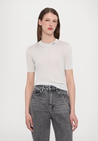 Top blanco de manga corta con canalé y escote redondo, con las letras "AIX" bordadas en negro. Combinado con jeans grises de cintura alta.