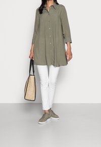Long green patterned button-up shirt, paired with white pants and grey sneakers. The model carries a textured beige and black handbag.