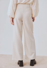 Pantalones de pierna ancha en tejido beige claro. Textura suave con un ajuste a medida y un único bolsillo trasero. Combinados con botas negras de tacón grueso.