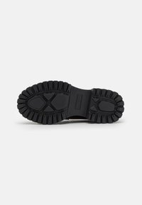 Semelle de chaussure en caoutchouc noir avec un motif de bande de roulement pour une meilleure adhérence, comportant des sections surélevées et une zone centrale lisse. La finition texturée renforce la durabilité.