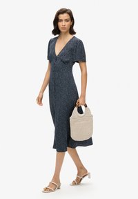 Granatowa sukienka midi z małymi białymi wzorami kwiatowymi, krótkimi bufkami i dekoltem w kształcie litery V. Modelka trzyma beżową plecioną torbę i ma na sobie beżowe sandały na paski.