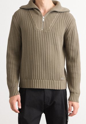 Mann trägt einen beigen, gerippten Strickpullover mit halbhohem Reißverschluss am Kragen, kombiniert mit schwarzen Hosen, steht vor einem schlichten Hintergrund.