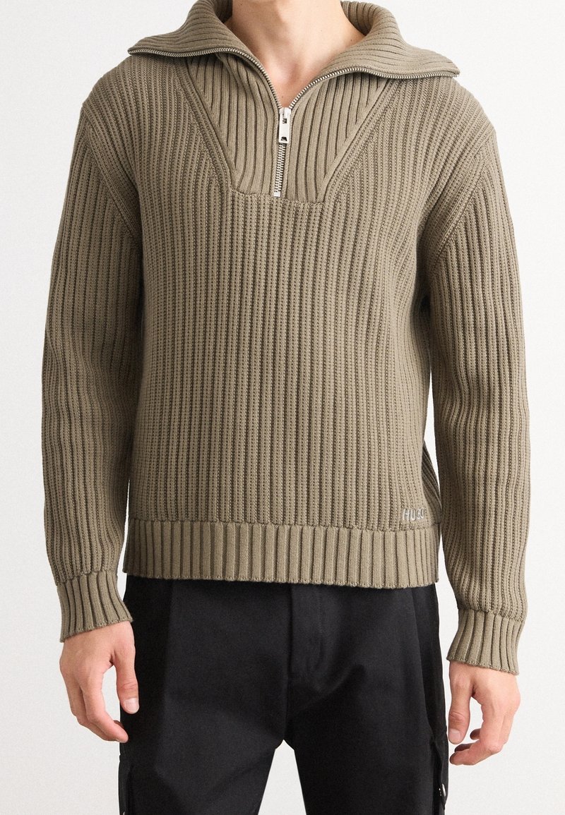 Homme portant un pull en tricot côtelé beige avec un col demi-fermeture éclair, assorti à un pantalon noir, debout devant un fond uni.