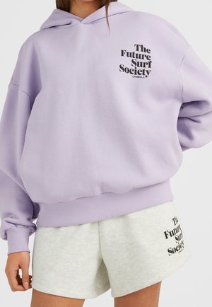 Person trägt lavendelfarbenen Hoodie und hellgraue Shorts, beide mit schwarzem Textlogo „The Future Surf Society“ auf der linken Seite.
