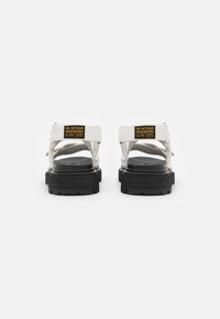 Witte sandalen met een gestructureerde zwarte zool, voorzien van verstelbare banden en een opvallend label op de achterkant. Strak, minimalistisch ontwerp.