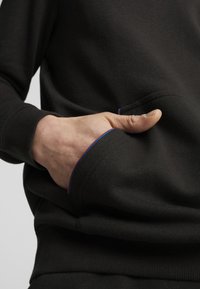 Černá mikina s kapucí a přední kapsou; má kontrastní modré švy podél kapsy a manžet; vyrobena z měkké, texturované látky.
