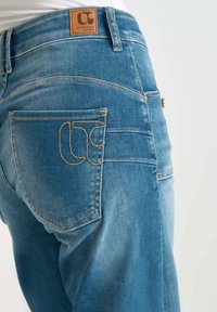Jeans in denim blu chiaro con una toppa in pelle marrone, cuciture gialle e un design unico del logo sulla tasca posteriore.