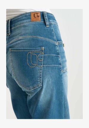 Light blue denim jeans featuring a brown leather patch, yellow stitching, and a unique logo design on the back pocket.