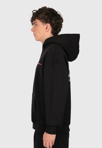 Junger Mann mit lockigem Haar, der einen schwarzen Kapuzenpullover mit hochgezogener Kapuze trägt, steht im Profil vor einem schlichten hellen Hintergrund.