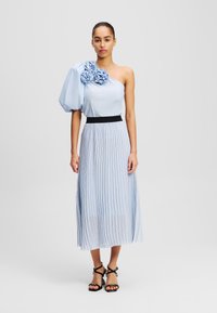 Robe asymétrique bleu clair avec un accent floral sur une épaule, jupe plissée et taille noire. Portée avec des talons noirs à lanières.