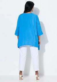 Tunica blu a maniche lunghe con scollatura rotonda, caratterizzata da una linea dritta e spacchi laterali, abbinata a pantaloni bianchi a cropped e scarpe con tacco.