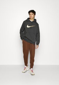 Tummanharmaa huppari, jossa on kultainen Nike-logo edessä, yhdistettynä ruskeisiin kapeneviin collegehousuihin. Asusteessa on joustavat hihansuut ja kiristysnauhat.