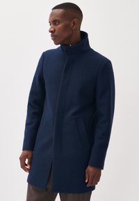 Abrigo de lana de color azul marino con un alto cuello, cierre de botones y bolsillos laterales. Textura suave con un diseño recto y ajustado.