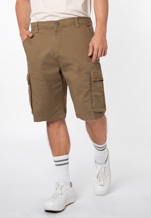 Man wearing brown cargo shorts, white socks with gray stripes, white sneakers, and a white t-shirt, standing with one hand in pocket.