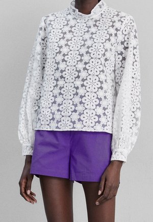 Person trägt eine weiße, lange Bluse aus floralem Spitzenstoff und lebendige lila Shorts, steht vor einem schlichten grauen Hintergrund.