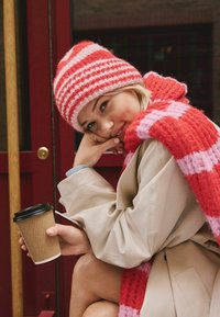 Knit hat in red and pink stripes, matching scarf in check pattern, cream coat, holding a brown paper coffee cup. Soft, textured materials.