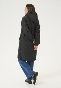 Manteau long matelassé noir avec capuche, présentant un motif en losanges et des manches longues. Porté avec un jean bleu évasé et des chaussures noires.