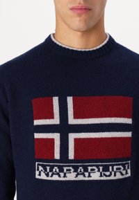 Dunkelblauer Strickpullover mit einem norwegischen Flaggenmotiv in Rot, Weiß und Blau, einem ribbed Kragen und dem Schriftzug "NAPAPIJRI" am unteren Ende.