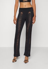 Kvinna iklädd svarta högmidjade stickade byxor med vertikala slitna paneler, tillsammans med svarta remmar med hög klack.