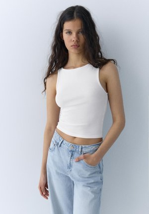 Young woman with long dark hair wearing a sleeveless white top and light blue jeans standing against a plain light wall.