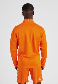 Heldere oranje sportkleding met een lang mouwtop met hoge kraag en bijpassende shorts. Ruwe textuur met logo-details op de shorts.
