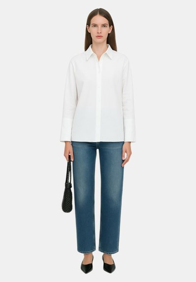 Camicia bianca con colletto, abbinata a jeans blu a gamba dritta. Il modello tiene una borsa intrecciata nera. Scarpe nere a punta completano il look.