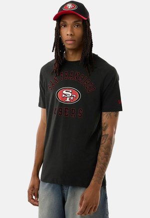 Mężczyzna z długimi włosami w czarnej czapce San Francisco 49ers i pasującej koszulce, stojący na jednolitym jasnym tle.