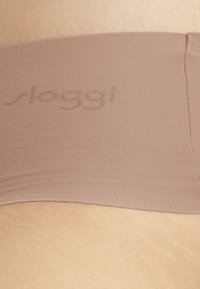 Brauner Bund aus dehnbarem Stoff mit glatter Textur, versehen mit einem dezenten Sloggi-Logo. Der darunterliegende Hautton ist leicht strukturiert.