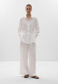Chemise en lin blanc avec taille à cordon de serrage, manches longues et bouton devant, associée à un pantalon large assorti et des tongs brunes.