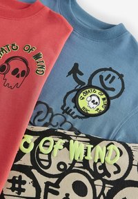 Divas sporta krekli: sarkans ar melnu skull grafiku un tekstu; zils ar apaļu uzlīmi, kas attēlo skull dizainu un melnām zīmēm.