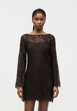 Γυναίκα που φοράει μαύρο φόρεμα με μακριά δαντελένια μανίκια και στρογγυλή λαιμόκοψη, στέκεται μπροστά σε απλό λευκό φόντο, κοιτώντας μπροστά.