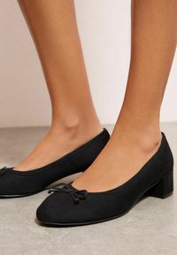 Černé baleríny z semiše s kulatou špičkou a malým blokovým podpatkem, zdobené dekorativní mašlí na přední části. Hladká textura.