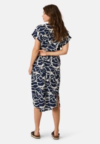 Marineblaues Kleid mit weißem abstraktem Muster, kurzen Ärmeln, seitlichen Schlitzen, weichem Stoff und knöchellangem Design. Modell trägt flache Sandalen.