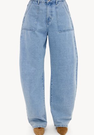 Jean bleu clair taille haute à jambes larges avec grandes poches plaquées devant et coutures visibles, porté avec des chaussures en daim marron à bout pointu.