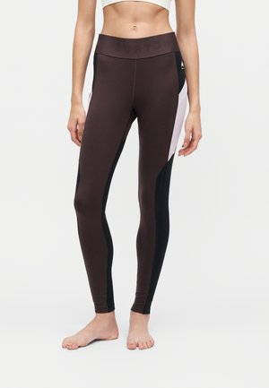 Femme portant un legging taille haute avec des blocs de couleur marron, noir et violet clair, debout pieds nus sur un fond blanc uni.