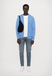 Κοντό φούτερ με φερμουάρ σε ανοιχτό μπλε χρώμα, λευκό t-shirt, ανοιχτό μπλε τζιν, μαύρη τσάντα ώμου και λευκά αθλητικά παπούτσια. Λείος ύφασμα, χαλαρός σχεδιασμός και casual στυλ.